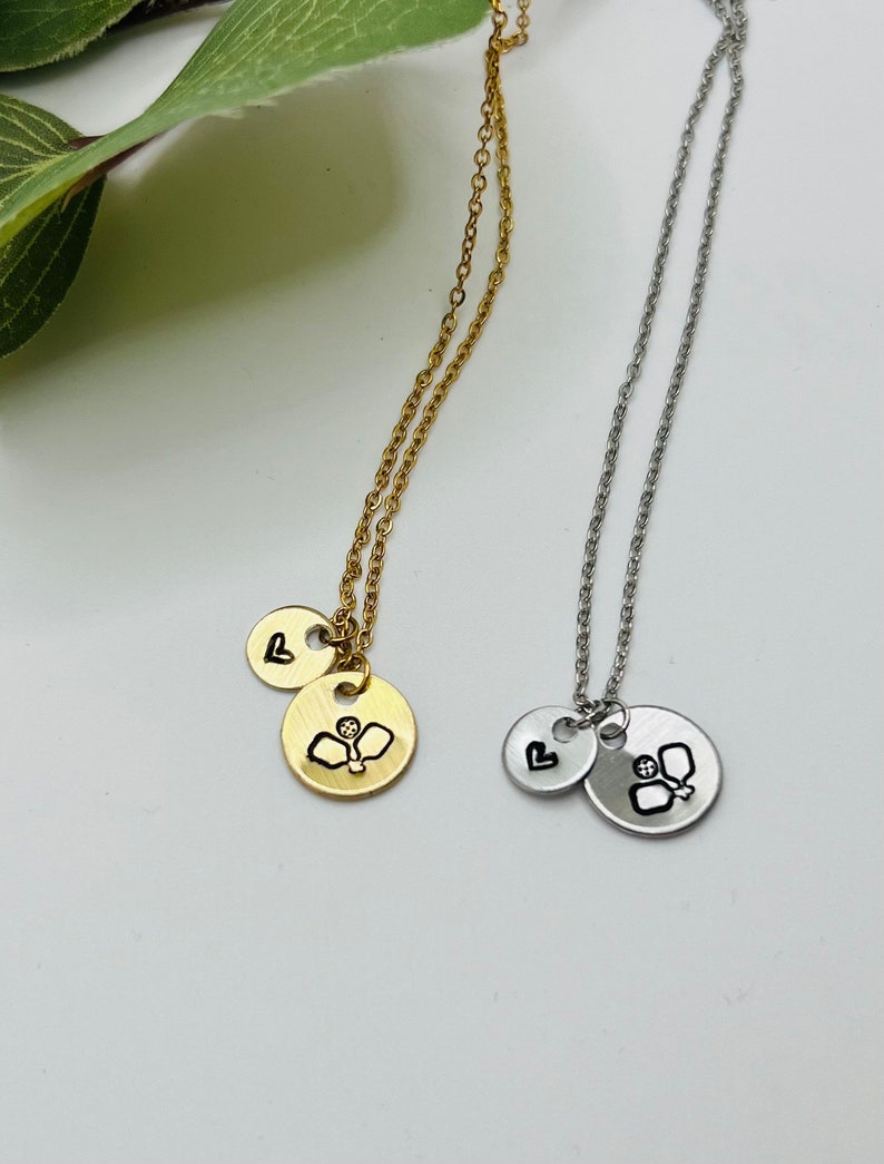 May include: Two necklaces with a heart charm and a charm with a paddle and ball design. One necklace is gold and the other is silver.