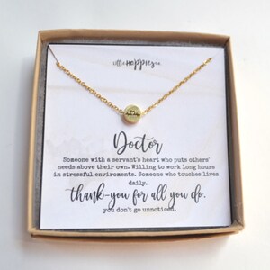 May include: Gold necklace with a small heart charm engraved with a heartbeat symbol. The necklace comes in a box with a card that reads "Doctor" and a message of appreciation for doctors.