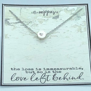 May include: A silver necklace with a small round pendant featuring a heart design. The necklace is displayed on a card with the text "the loss is immeasurable, but so is the love left behind."