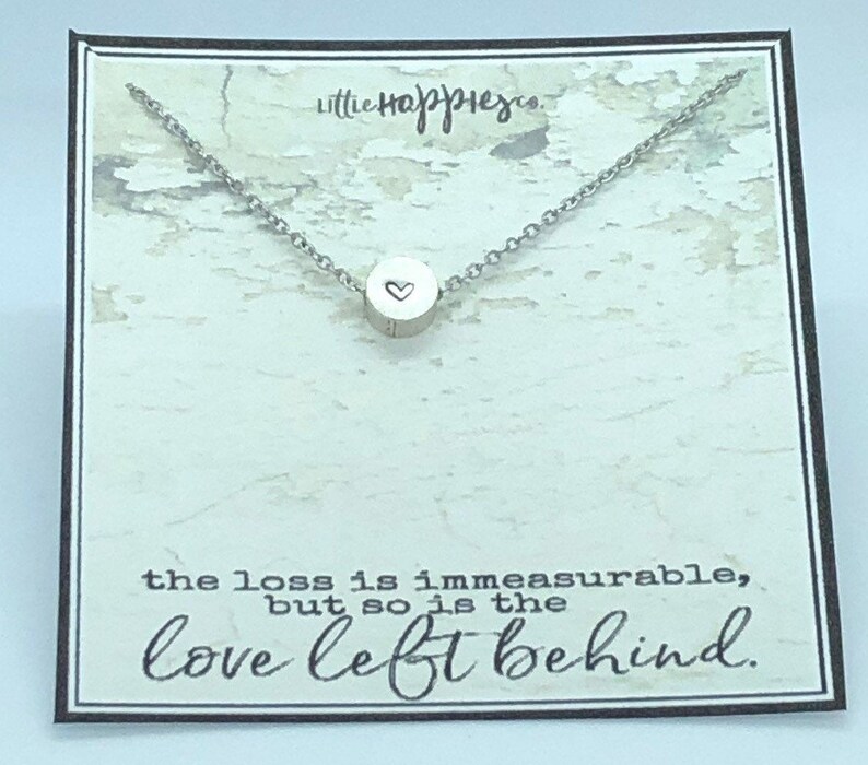 May include: A silver necklace with a small round pendant featuring a heart design. The necklace is displayed on a card with the text "the loss is immeasurable, but so is the love left behind."