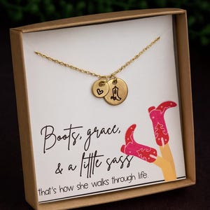 May include: A gold-colored necklace with two charms, one heart-shaped and the other a cowboy boot. The necklace is presented in a gift box with the text "Boots, grace, & a little sass" and an illustration of pink cowboy boots.