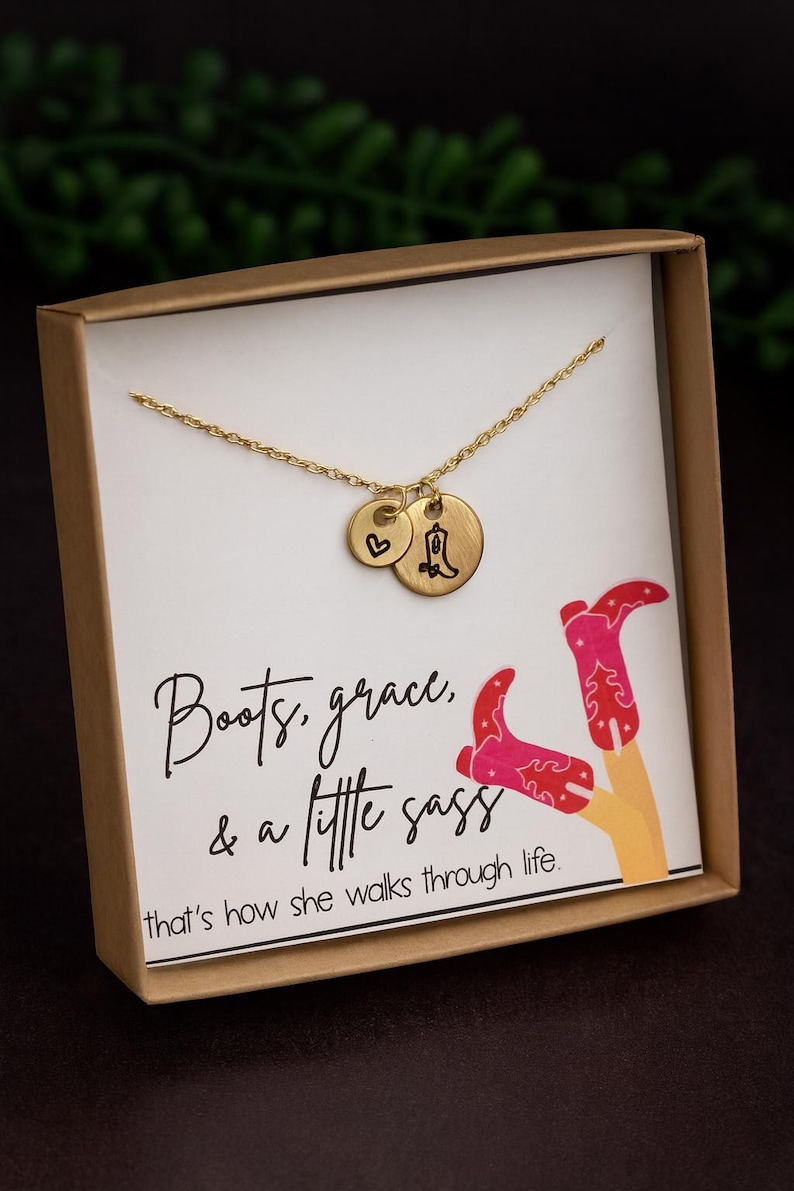 May include: A gold-colored necklace with two charms, one heart-shaped and the other a cowboy boot. The necklace is presented in a gift box with the text "Boots, grace, & a little sass" and an illustration of pink cowboy boots.