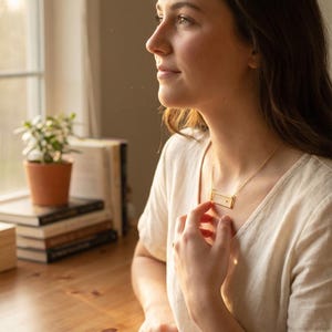 May include: A gold-toned necklace featuring a horizontal rectangular pendant with a small, centered accent stone. The pendant is suspended from a delicate, thin gold-toned chain that rests against the collarbone area of the wearer.