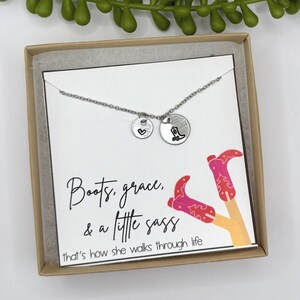 May include: A silver necklace with two round charms, one with a heart and the other with a cowboy boot. The necklace is displayed in a gift box with a card that reads "Boots, grace, & a little sass".