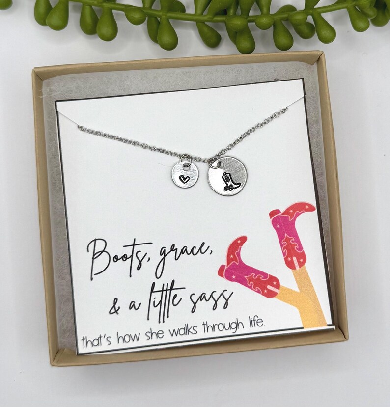 May include: A silver necklace with two round charms, one with a heart and the other with a cowboy boot. The necklace is displayed in a gift box with a card that reads "Boots, grace, & a little sass".