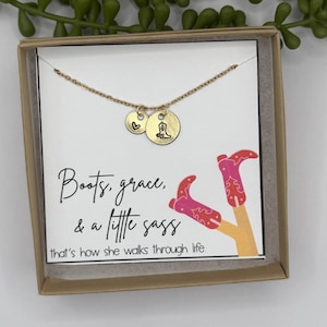 May include: A gold-colored necklace with two charms, one heart-shaped and one round with a boot design. The necklace is presented in a gift box with the text "Boots, grace, & a little sass" and a drawing of pink cowboy boots.