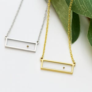 May include: Two necklaces with thin metal chains and rectangular open-frame pendants. One necklace is silver-toned and the other is gold-toned. Each pendant features a single small, dark circular dot positioned toward the right side of the frame.