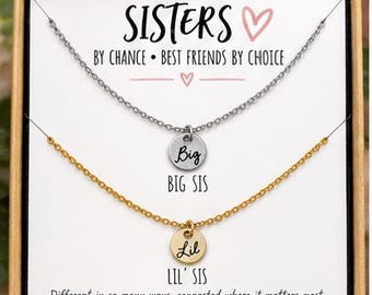 May include: Two necklaces in a gift box. One silver-tone chain has a round pendant engraved with Big, and one gold-tone chain has a round pendant engraved with Lil. The card reads SISTERS BY CHANCE • BEST FRIENDS BY CHOICE, BIG SIS, LIL' SIS.