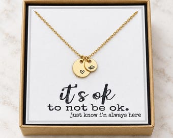 May include: A gold-toned necklace with two circular charms, one featuring a small heart and the other a wing icon. The necklace is displayed on a white card inside a brown box, with the text "it's ok to not be ok. just know i'm always here."