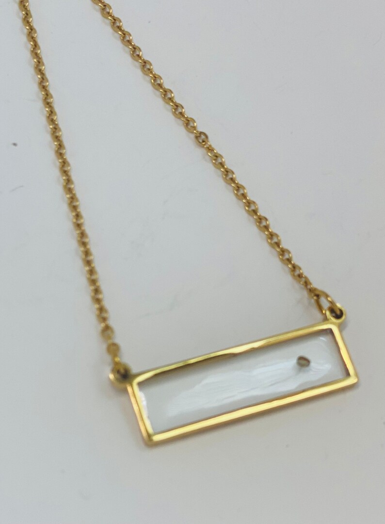 Pu&ograve; includere: Una collana con catena dorata e un ciondolo rettangolare. Il ciondolo &egrave; trasparente e contiene un piccolo oggetto argentato all'interno.