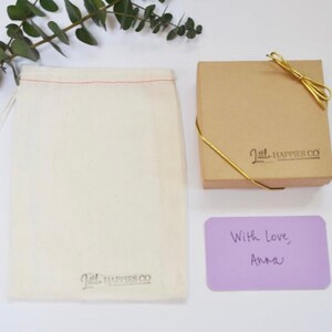 May include: A small, white cotton drawstring bag with the text "Little Happies Co." printed on the bottom. Next to the bag is a small, brown cardboard box with a gold ribbon and the text "Little Happies Co." printed on the top. A purple card with the text "With love, Anna" is in front of the box.