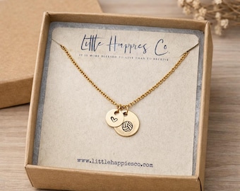 May include: A gold-toned chain necklace featuring two small circular charms, one engraved with a heart and the other with a volleyball. The necklace is presented on a branded card inside a brown cardboard box with the text Little Happies Co.