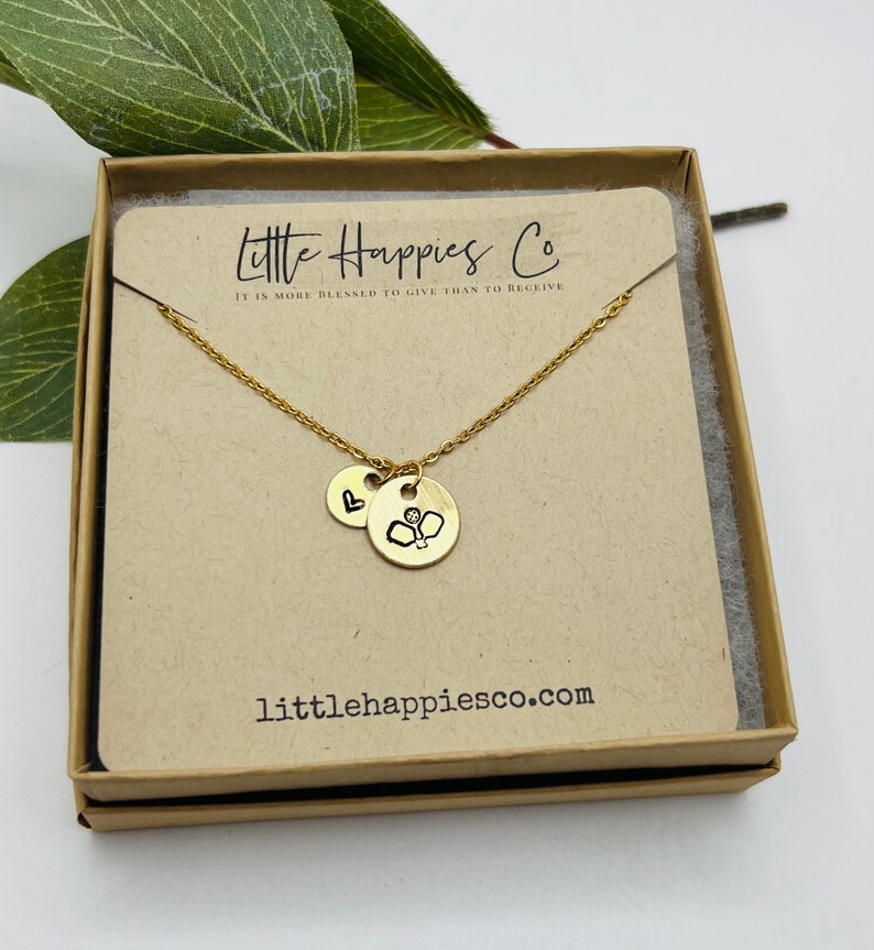May include: Gold necklace with two charms, one with a heart and the other with a boxing glove. The necklace is in a brown box with the text "Little Happies Co. It is more blessed to give than to receive" and "littlehappiesco.com".