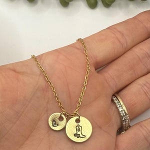 May include: Gold-colored necklace with two circular pendants. One pendant features the letter "L", the other a cowboy boot. The necklace is held in a hand, with a ring visible on a finger. The background is white with a green plant.