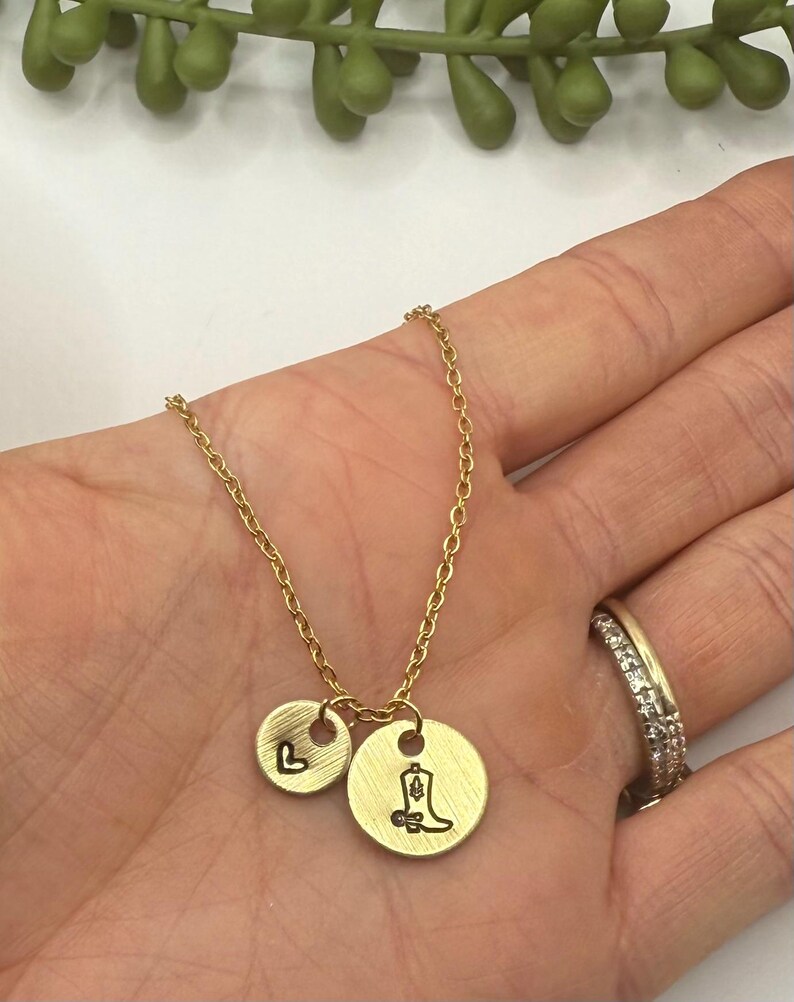 May include: Gold-colored necklace with two circular pendants. One pendant features the letter "L", the other a cowboy boot. The necklace is held in a hand, with a ring visible on a finger. The background is white with a green plant.