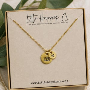 May include: A gold-toned necklace featuring two circular charms, one with a camera icon and one with a heart symbol, displayed on a beige card printed with Little Happies Co and the text IT IS MORE BLESSED TO GIVE THAN TO RECEIVE and the website URL.