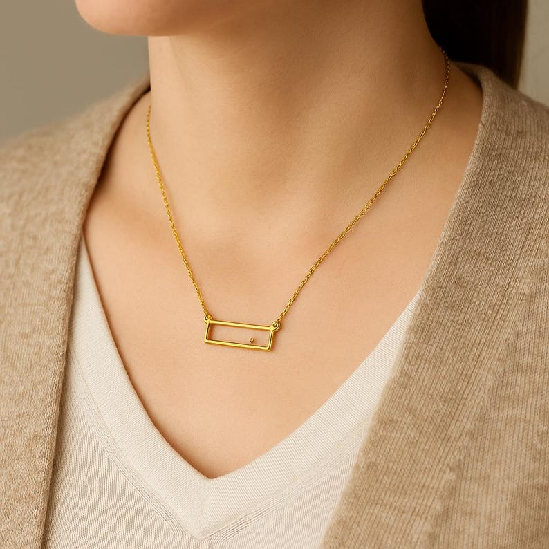 May include: A gold-toned necklace featuring a delicate chain and a rectangular pendant frame. The pendant is an open, thin gold rectangle with a single small, round, neutral-colored stone set along the bottom edge of the frame, suspended from a chain.