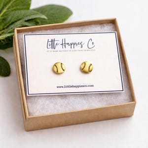 May include: A pair of gold-colored baseball stud earrings presented in a small, open cardboard box. The earrings are on a white card with the text "Little Happies Co" and "IT IS MORE BLESSED TO GIVE THAN TO RECEIVE". The website address www.littlehappiesco.com is also visible.