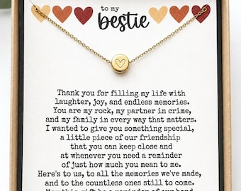 Peut inclure: Un collier doré avec un pendentif en forme de cœur est présenté dans un coffret cadeau. La boîte contient une carte avec le texte "to my bestie" et un message. La carte est ornée de cœurs colorés.