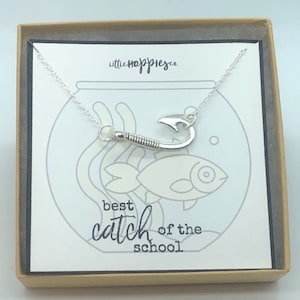May include: A silver necklace with a fish hook charm in a box with a white background. The box has the text "little happies co." and "best catch of the school" printed on it.