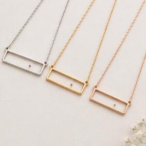May include: Three necklaces featuring a thin rectangular frame pendant suspended from a delicate chain. The necklaces are presented in silver, gold, and rose gold tones, each with a single small dot centered within the open rectangular frame.