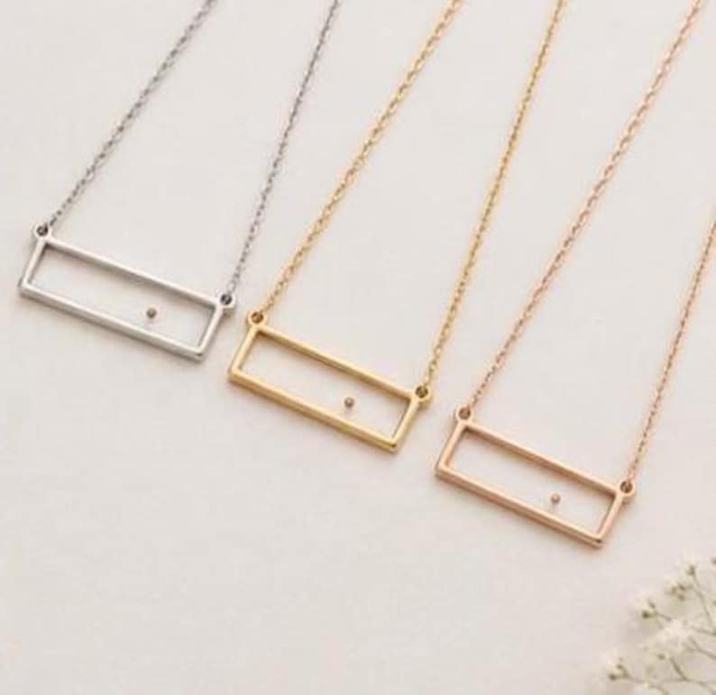 May include: Three necklaces featuring a thin rectangular frame pendant suspended from a delicate chain. The necklaces are presented in silver, gold, and rose gold tones, each with a single small dot centered within the open rectangular frame.