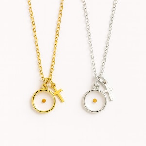 May include: Two necklaces with delicate chains. One is gold-toned, the other silver-toned. Each features a pendant with a small mustard seed inside a circle, and a small cross charm. The background is white.
