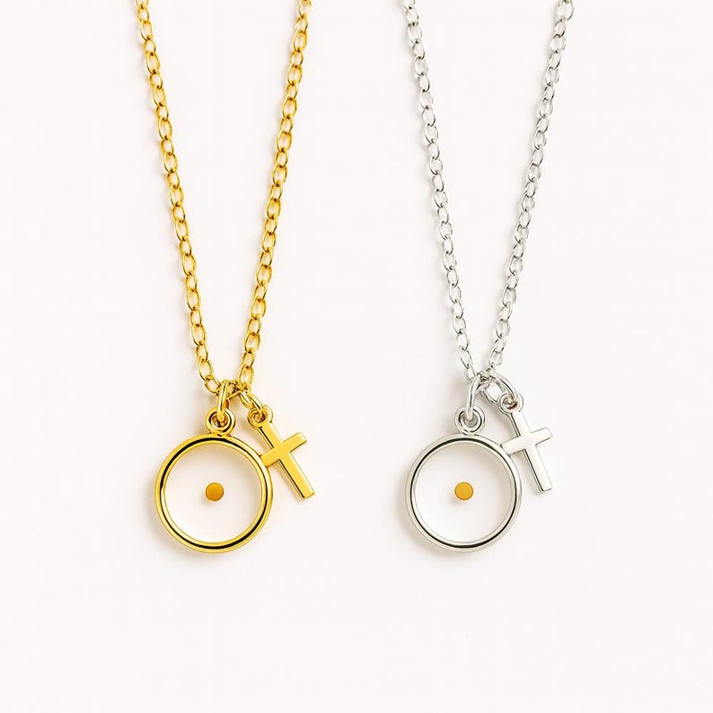 May include: Two necklaces with delicate chains. One is gold-toned, the other silver-toned. Each features a pendant with a small mustard seed inside a circle, and a small cross charm. The background is white.