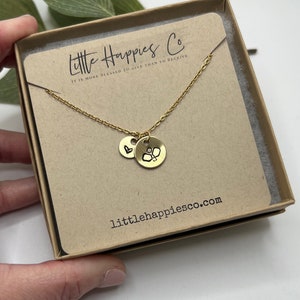 May include: Gold necklace featuring a small heart charm and a larger charm with a pickleball design. The necklace is presented in a small box with the text "Little Happies Co. It is more blessed to give than to receive" and the website "littlehappiesco.com".
