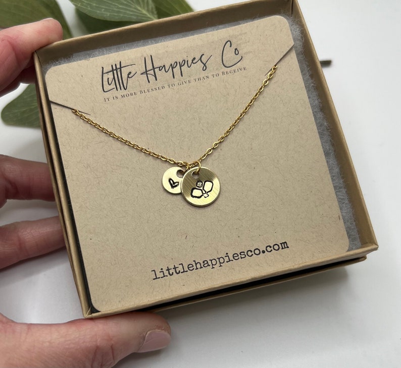 May include: Gold necklace featuring a small heart charm and a larger charm with a pickleball design. The necklace is presented in a small box with the text "Little Happies Co. It is more blessed to give than to receive" and the website "littlehappiesco.com".