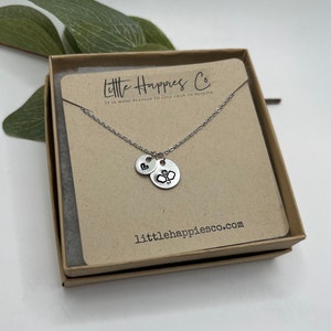 May include: A silver necklace with two charms, one with a heart and the other with a badminton racket. The necklace is in a brown box with the text "Little Happies Co. It is more blessed to give than to receive." and "littlehappiesco.com".