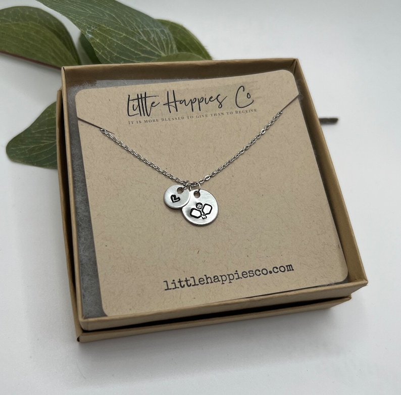May include: A silver necklace with two charms, one with a heart and the other with a badminton racket. The necklace is in a brown box with the text "Little Happies Co. It is more blessed to give than to receive." and "littlehappiesco.com".