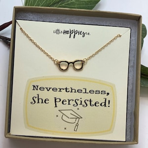 May include: A gold necklace with a pair of glasses charm in a box with a yellow background. The box has the text "Nevertheles, She Persisted!" and a graduation cap graphic.