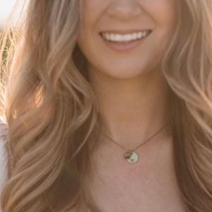 May include: A woman with long, wavy blonde hair smiles, wearing a delicate necklace with a small, round pendant. The necklace is centered on the chest. The background is blurred with warm, golden light.