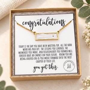 May include: A gold-toned bar necklace with a rectangular white iridescent stone, presented in a cardboard gift box on a card that reads congratulations, today is the day you have been waiting for, and you got this with a circular logo.