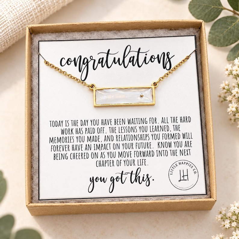 May include: A gold-toned bar necklace with a rectangular white iridescent stone, presented in a cardboard gift box on a card that reads congratulations, today is the day you have been waiting for, and you got this with a circular logo.