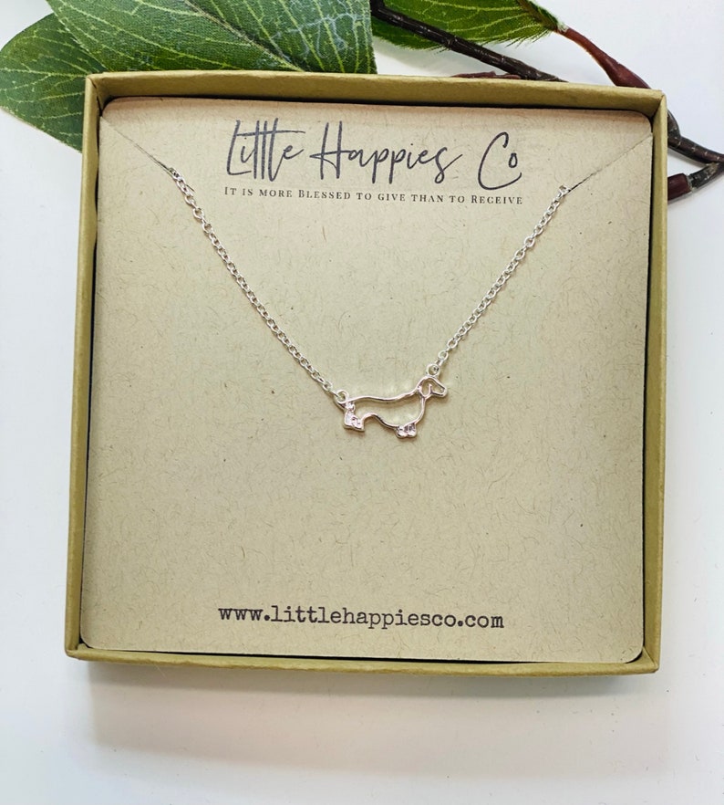 Peut inclure: Un collier en argent en forme de dachshund dans une petite bo&icirc;te avec le texte "Little Happies Co. It is more blessed to give than to receive" et le site web "www.littlehappiesco.com".