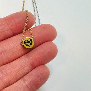 K&ouml;nnte beinhalten: Goldfarbene Halskette mit einem kleinen, runden Anh&auml;nger. Der Anh&auml;nger hat einen gelben Hintergrund mit einem schwarzen herzf&ouml;rmigen Design, das mit kleinen schwarzen Punkten gef&uuml;llt ist. Die Halskette wird in einer Hand vor wei&szlig;em Hintergrund gehalten.