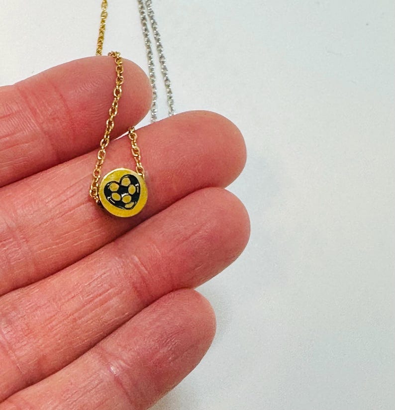 K&ouml;nnte beinhalten: Goldfarbene Halskette mit einem kleinen, runden Anh&auml;nger. Der Anh&auml;nger hat einen gelben Hintergrund mit einem schwarzen herzf&ouml;rmigen Design, das mit kleinen schwarzen Punkten gef&uuml;llt ist. Die Halskette wird in einer Hand vor wei&szlig;em Hintergrund gehalten.