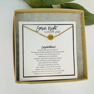 K&ouml;nnte beinhalten: Eine goldfarbene Halskette mit rundem Anh&auml;nger, pr&auml;sentiert in einer Geschenkbox. Der Anh&auml;nger ist mit einem Design versehen. Die Karte in der Box tr&auml;gt die Aufschrift "Senior Night custom year" und "Herzlichen Gl&uuml;ckwunsch!" mit einer Nachricht.
