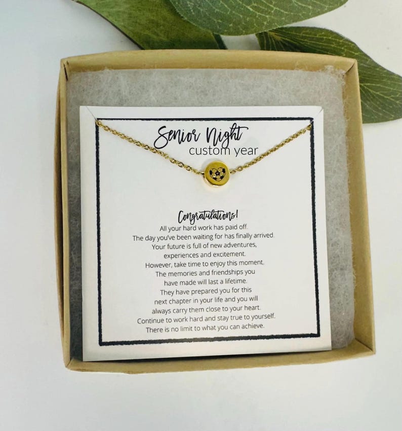 K&ouml;nnte beinhalten: Eine goldfarbene Halskette mit rundem Anh&auml;nger, pr&auml;sentiert in einer Geschenkbox. Der Anh&auml;nger ist mit einem Design versehen. Die Karte in der Box tr&auml;gt die Aufschrift "Senior Night custom year" und "Herzlichen Gl&uuml;ckwunsch!" mit einer Nachricht.