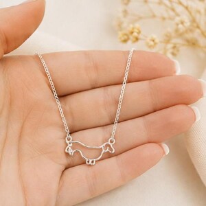 May include: A silver-toned necklace featuring a delicate, continuous wire-outline pendant shaped like a dachshund dog. The pendant is suspended from a thin, matching silver-toned chain, creating a simple and clean aesthetic for this piece of jewellery.