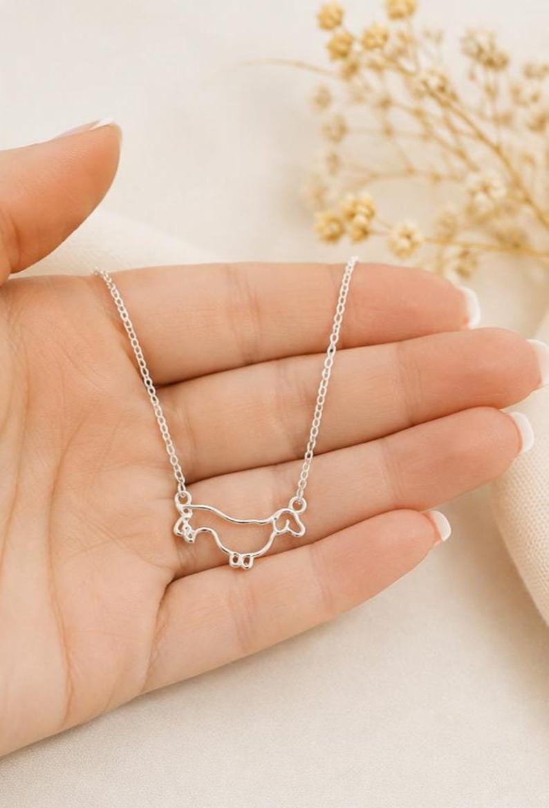 May include: A silver-toned necklace featuring a delicate, continuous wire-outline pendant shaped like a dachshund dog. The pendant is suspended from a thin, matching silver-toned chain, creating a simple and clean aesthetic for this piece of jewellery.