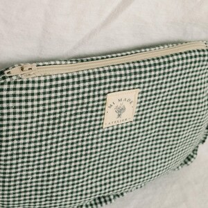 May include: A green and white gingham pattern pouch with a beige zip. The pouch features a rectangular label that reads "MI MADE - ATELIER" with a floral design. The pouch is on a white surface.