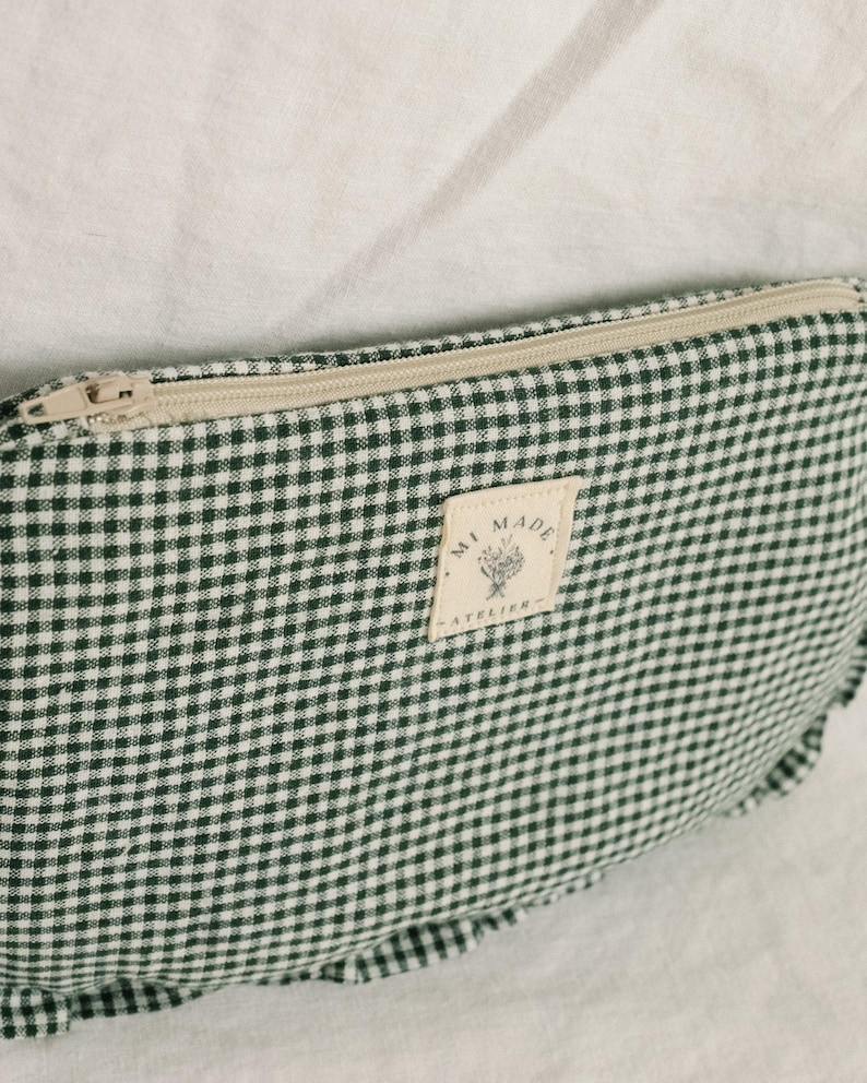 May include: A green and white gingham pattern pouch with a beige zip. The pouch features a rectangular label that reads "MI MADE - ATELIER" with a floral design. The pouch is on a white surface.