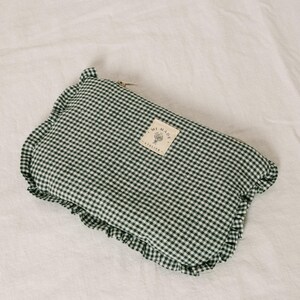 May include: A green and white gingham patterned pouch with a ruffled edge. The pouch has a small rectangular tag with text. The pouch is laying on a white surface.