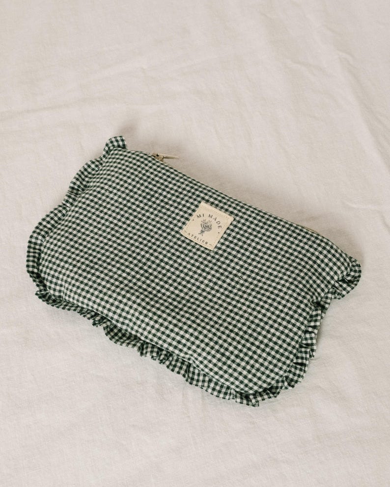 May include: A green and white gingham patterned pouch with a ruffled edge. The pouch has a small rectangular tag with text. The pouch is laying on a white surface.