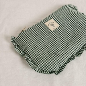 May include: A folded green and white gingham fabric pouch with a ruffled edge. A small tag with text and a floral design is attached. The pouch is closed with a metal clasp. The fabric is a classic checkered pattern.