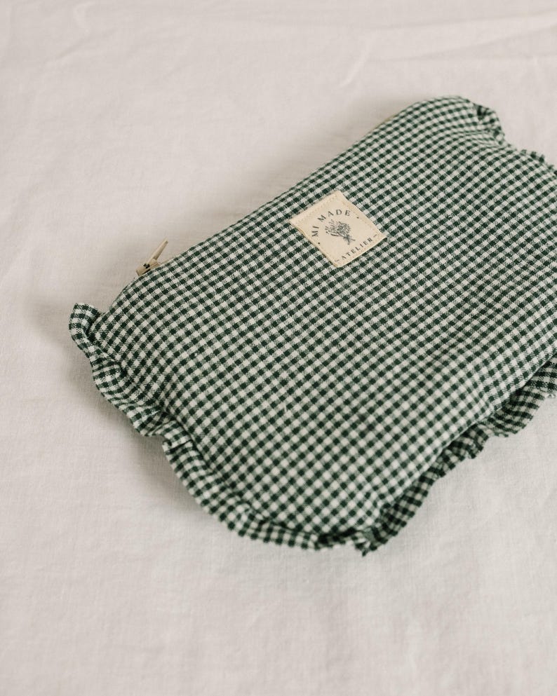 May include: A folded green and white gingham fabric pouch with a ruffled edge. A small tag with text and a floral design is attached. The pouch is closed with a metal clasp. The fabric is a classic checkered pattern.