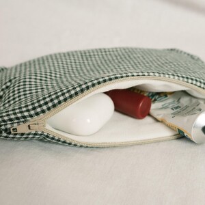 May include: A green and white gingham cosmetic bag with a beige zip, open to reveal a white bar of soap, a red tube, and a tube of cream. The bag is on a white surface.